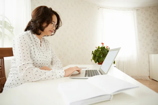 Side view of serious mature woman in glasses using wireless Internet connection on laptop, shopping online or reading news, sitting at the white table against home interior background. Selective focus