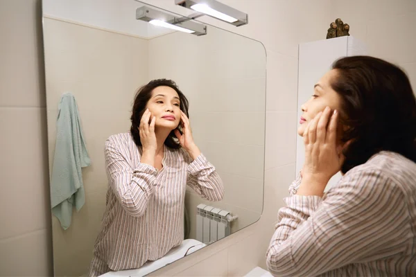 Mature brunette female applying make-up while doing morning routine procedures. Portrait of beautiful middle-aged woman wearing pajamas, looking at herself in the mirror. Beauty and health concept