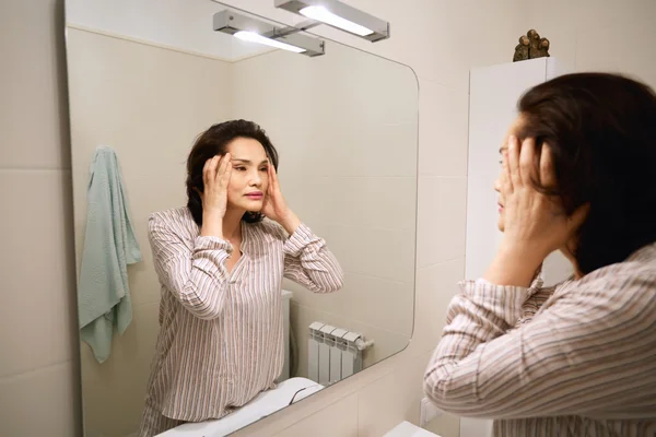Portrait of attractive middle-aged Asian female examining her skin for wrinkles, holding her face, looking at her reflection in the mirror. Adult brunette woman applying anti-aging cream or lotion