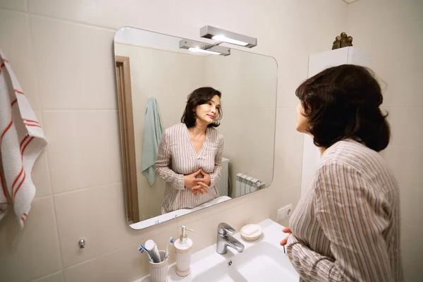 Beauty and health concept. Picture of attractive brunette Asian senior woman wearing pajamas, looking and smiling at herself in the mirror while doing night routine procedures before going to bed