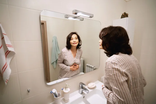 Portrait of beautiful adult woman of middle age looking at herself in the mirror and smiling. Elderly female applying anti-aging cream or make-up in the bathroom before going shopping in the morning