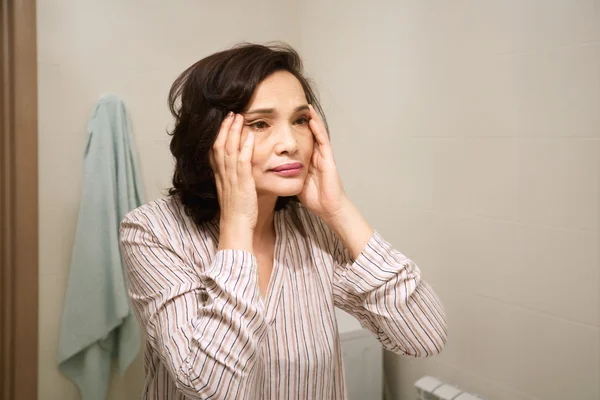 Portrait of middle-aged woman's reflection. Beautiful brunette elderly female wearing pajamas, applying anti-aging cream, looking for ageing signs, standing in front of the mirror in the bathroom.