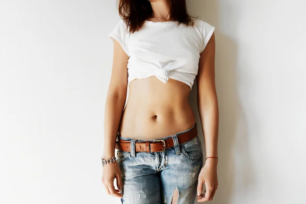 Woman with perfect slim and tanned body wearing denim pants. Young female model standing in fashion pose showing her nude belly while posing against white copy space wall for your advertising content