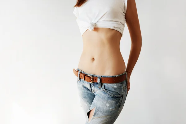 Close up of flat abdomen of young woman in ripped jeans. Cropped portrait of brunette with young healthy skin and naked belly. Young female with slim perfect body wearing trendy clothing indoor