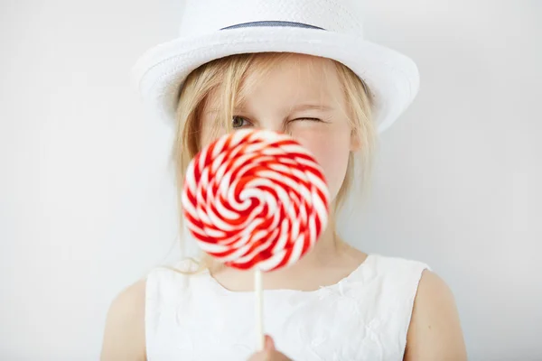 Close up portrait of beautiful little female child in white hat holding huge spiral lollipop, having fun while enjoying sweet candy with happy expression, against white concrete wall. Selective focus