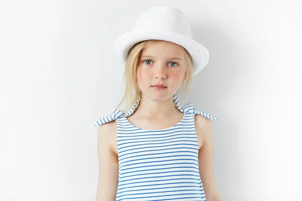 People and lifestyle concept. Close up of happy little female model posing against white concrete wall, wearing white hat and striped dress. Adorable blonde preschool girl looking at the camera