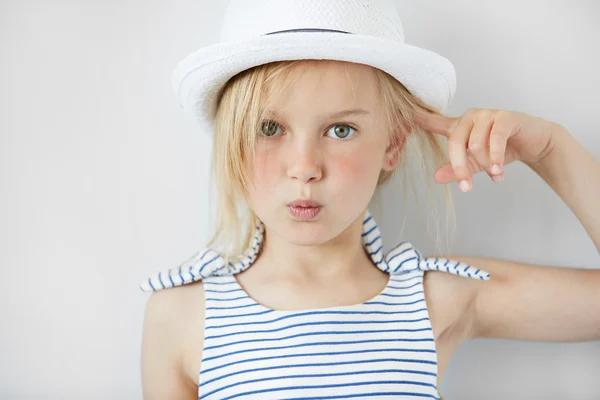 Close up shot of  annoyed and angry girl wearing white hat and striped dress, holding index finger at her head with \'are you crazy\' gesture. Isolated portrait of little Caucasian 5-year old child