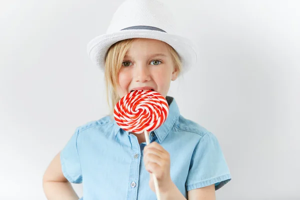 Adorable Caucasian little girl licking big colorful spiral lollipop. Cute preschool child wearing fashionable clothes looking at the camera while enjoying sweet candy. People and lifestyle concept