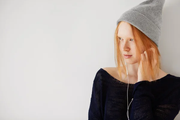 Redhead teenage girl wearing hipster cap listening to her favorite music with earphones. Pretty schoolgirl in headphones dancing against white background with copy space for your promotional content