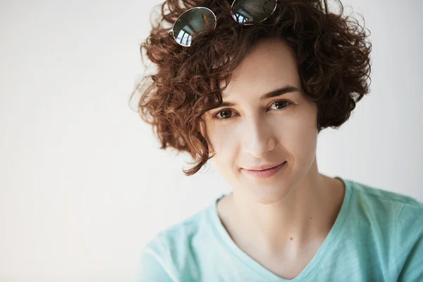 Close up portrait of beautiful young woman with short curly hair smiling at the camera. Happy female with sunglasses on her head looking with confident expression on her face, achieving life goals