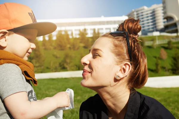Portrait of happy redhead mother playing with her two-year old son wearing stylish cap during nice walk in the park on sunny day. Young female having fun with her baby boy. Family relations concept