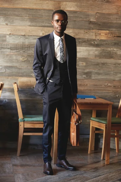 Full-length portrait of handsome African businessman wearing elegant formal suit, holding leather bag in his hands, posing against cozy cafe interior, looking at the camera with confident expression