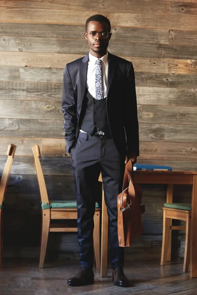 Success and career concept. Full-length shot of successful African entrepreneur in formal wear, holding briefcase, looking at the camera with serious expression, standing against coffee shop interior