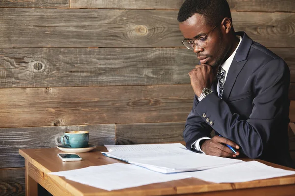 Handsome young African entrepreneur reading a contract before signing it, resting his elbow on the wooden table at a restaurant, sitting with confident concentrated look against wooden wall background