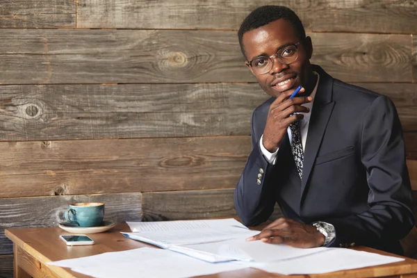 Happy African student in formal suit and glasses looking and smiling at the camera, holding a hand on his chin, doing homework while sitting at the wooden table with cell phone and a cup of coffee