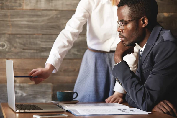 African CEO in formal wear sitting at the table in front of laptop, listening attentively to presentation of a business project by his redhead Caucasian assistant, who points at the screen with a pen