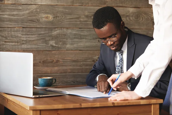 Focused African American businessman checking papers with his personal assistant in white shirt. Young female colleague points at lines in documents, explaining details in a business contract.