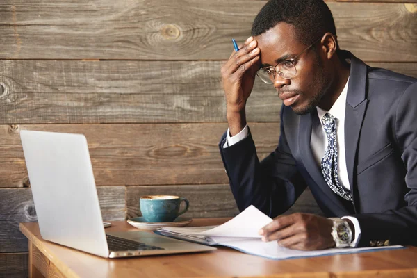 Worried African businessman in official suit checking information in laptop and papers in cosy cafe. He\'s making hard decision, looking focused, confused and puzzled with hand rested on his forehead.