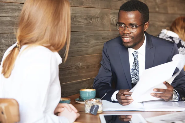 Human resource concept. Portrait of African American HR director wearing formal suit interviewing redhead Caucasian female, checking her resume during a meeting at a coffee shop. Business and career