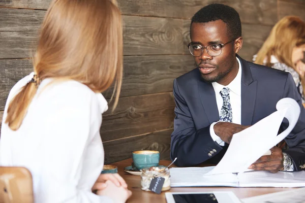 African boss conducting an interview with redhead Caucasian woman, holding job application and listening attentively to candidate. Two office workers discussing a project during lunch break at a cafe