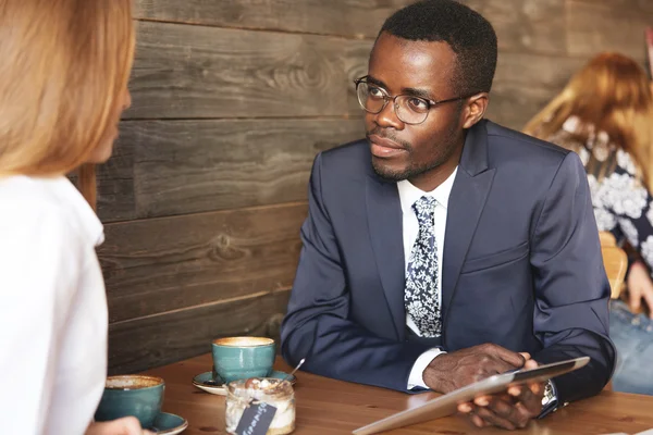 Team work: two corporate workers in formal wear sitting together at the table and discussing business plans. African man using digital tablet during a meeting with his Caucasian female colleague