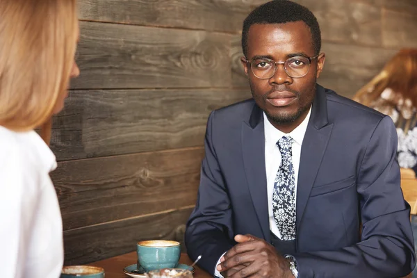 Portrait of confident attractive African businessman in glasses and formal suit looking at the camera, sitting at the table at a coffee while having business lunch with his redhead Caucasian partner