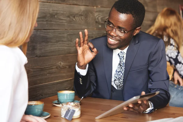 We already have great results! African American businessman holding digital tablet, gesturing, watching presentation, made by his redhead Caucasian colleague, with pleased and satisfied expression