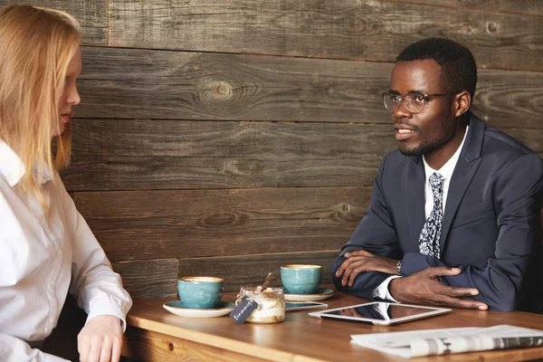 Young African entrepreneur in round glasses sitting at business lunch in a cafe and drinking coffee with female coworker. Businessman looking thoughtfully ahead, listening with arms crossed on table.