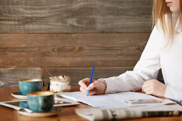 Cropped shot of young redhead female employee working at a coffee shop. Close up of student girl\'s hands holding a pen and writing down information in her copybook while doing homework at a cafe