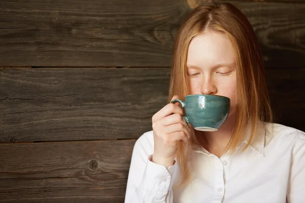 Young beautiful lady drinking tasty coffee on wooden background. Blond Caucasian girl in office dress having lunch in cafe sitting alone. She looks natural and relaxed in her beauty with eyes down.