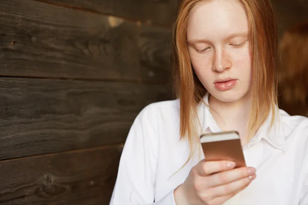 Portrait of young blond Caucasian girl with ivory skin and freckles. Business lady looking down at her smartphone, checking news or chatting in social networks. Office worker sitting in cafe