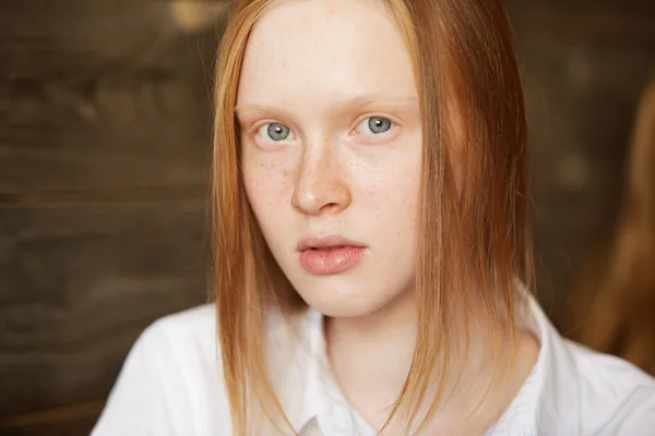 Close up shot of young female with blue eyes and freckled fresh skin looking at the camera with serious expression. Redhead Caucasian student girl relaxing at a cafe after lectures at university