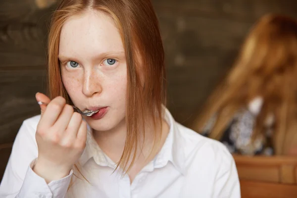 Close up shot of teenager eating dessert with a spoon, looking at the camera. Young Caucasian student girl with red hair and freckles wearing white shirt, enjoying delicious cake during lunch break