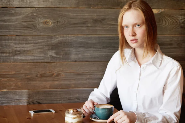 Redhead teenage girl in white shirt having morning cappuccino while sitting at a coffee shop before going to lectures at university, using cell phone with blank copy space screen for your text