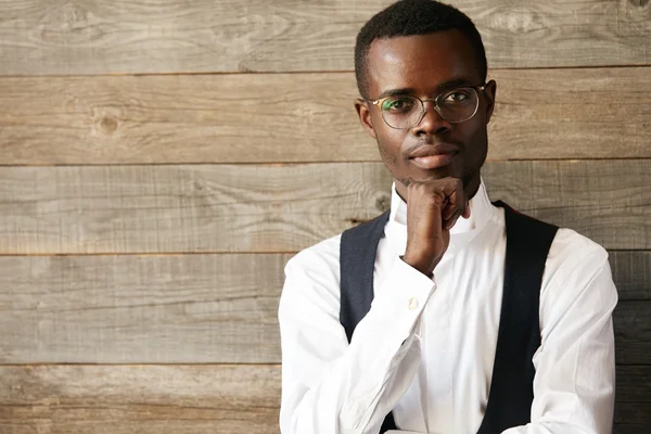 Close up isolated view of serious handsome young African male in white shirt and waistcoat looking with thoughtful expression at the camera. Lifestyle and people concept. Human expressions, emotions
