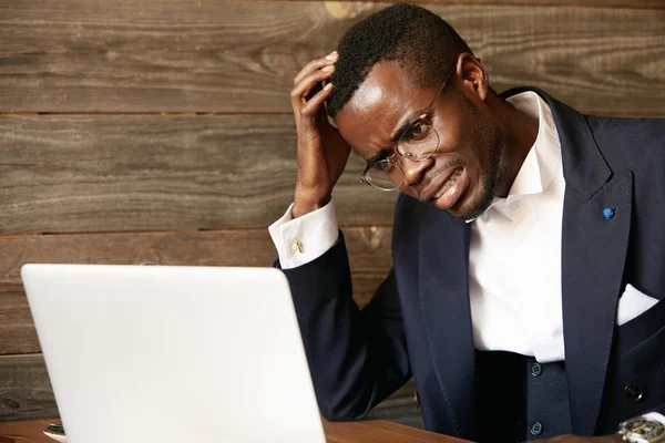 Dark-skinned businessman in formal suit and glasses having stress while working on laptop at the cafe, looking at the screen with stressed and frustrated expression, resting his elbow on the table