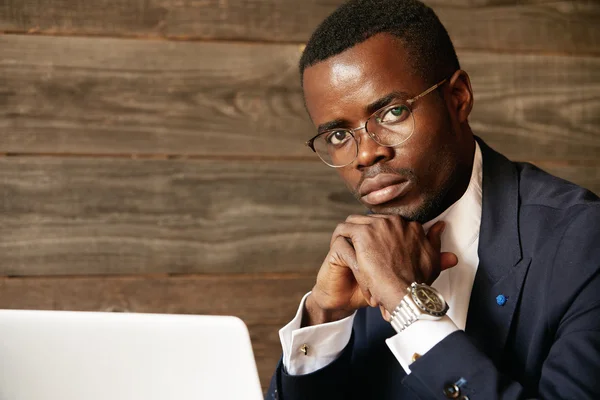 Young African office worker wearing elegant formal wear and glasses while using wireless Internet connection for distant work. Serious black man sitting with crossed hands. looking at the camera