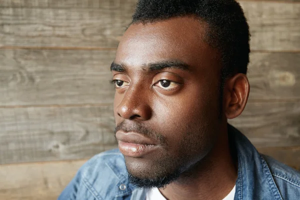 Highly-detailed profile of handsome young African man wearing denim jacket over white T-shirt looking with thoughtful and dreaming expression, making plans for the day, against wooden wall background