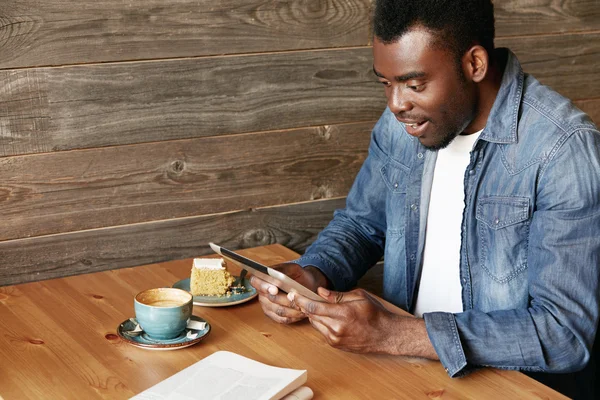 African man holding digital tablet with both hands, looking with complete disbelief, with mouth open, surprised with big sale prices while shopping online. Black freelancer sitting at a coffee shop