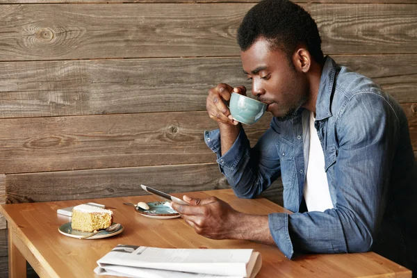 Handsome black male in casual wear checking email or reading world news on digital tablet computer drinking morning cappuccino, sitting at the wooden table at a cozy cafe. Connection and technology