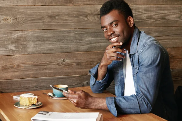 Young African student or freelancer wearing denim jacket looking and smiling at the camera, holding chin, resting his elbow on the wooden table, eating cake and drinking coffee, using digital tablet