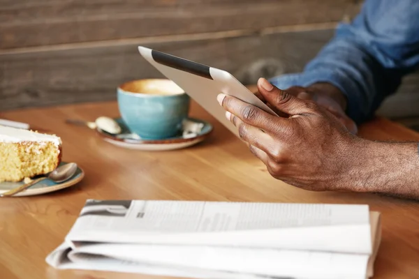 Close up view of African man\'s hands holding digital tablet, typing a message via social networks. Young black freelancer using electronic device for distance work while sitting at a cozy cafe.