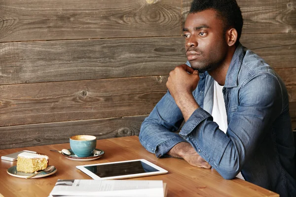 African man looking into the distance with thoughtful and dreamy expression, making plans for future, holding a hand on chin, using digital tablet with copy space screen for your advertising content