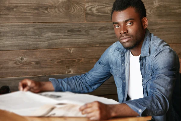 Selective focus. Young African student sitting at the wooden table at a coffee shop while waiting for his friend who is late. Black man in denim clothes looking at the camera with serious expression