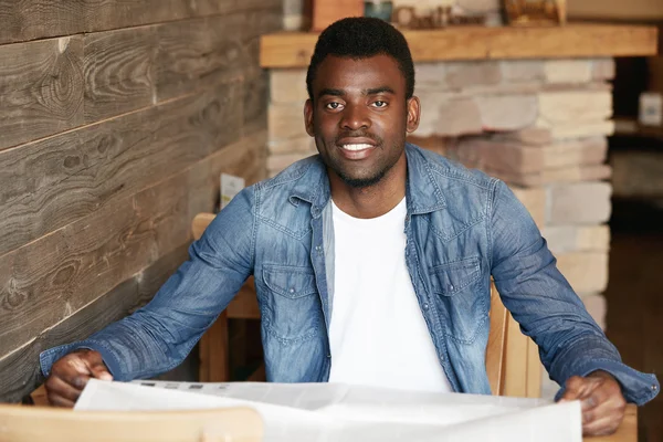 Handsome black man in denim jacket over white T-shirt looking at the camera with happy ultrawhite smile, enjoying leisure time at a coffee shop, reading tabloid. Human face expressions and emotions