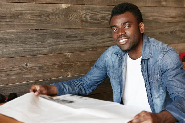 Attractive young African office worker in denim jacket reading tabloid while sitting at the wooden table at a restaurant during lunch break. Handsome black student relaxing at a student cafe