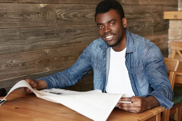 Lifestyle and people concept. Portrait of handsome young African American student in denim clothes, looking through morning newspaper while waiting for friends at a cozy cafe with wooden walls