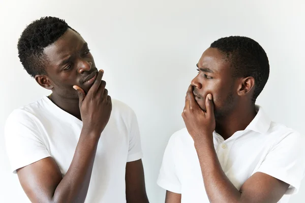 Portrait of two cautious and thoughtful black guys wearing white T-shirts looking at each other with skeptical and suspicious expression, holding hands on chin. Human face expressions and emotions