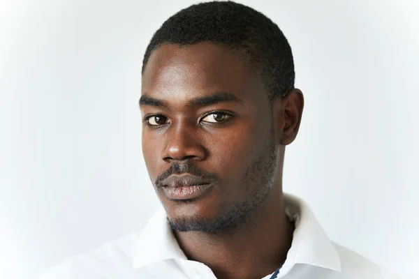 Half profile shot of attractive young African man with moustache and beard in casual clothes, looking at the camera with serious confident expression, posing against white concrete wall background