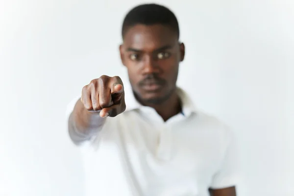 Isolated studio portrait of young dark-skinned student in white shirt, pointing at the viewer, looking at the camera with serious expression. Selective focus on the index finger. Body language